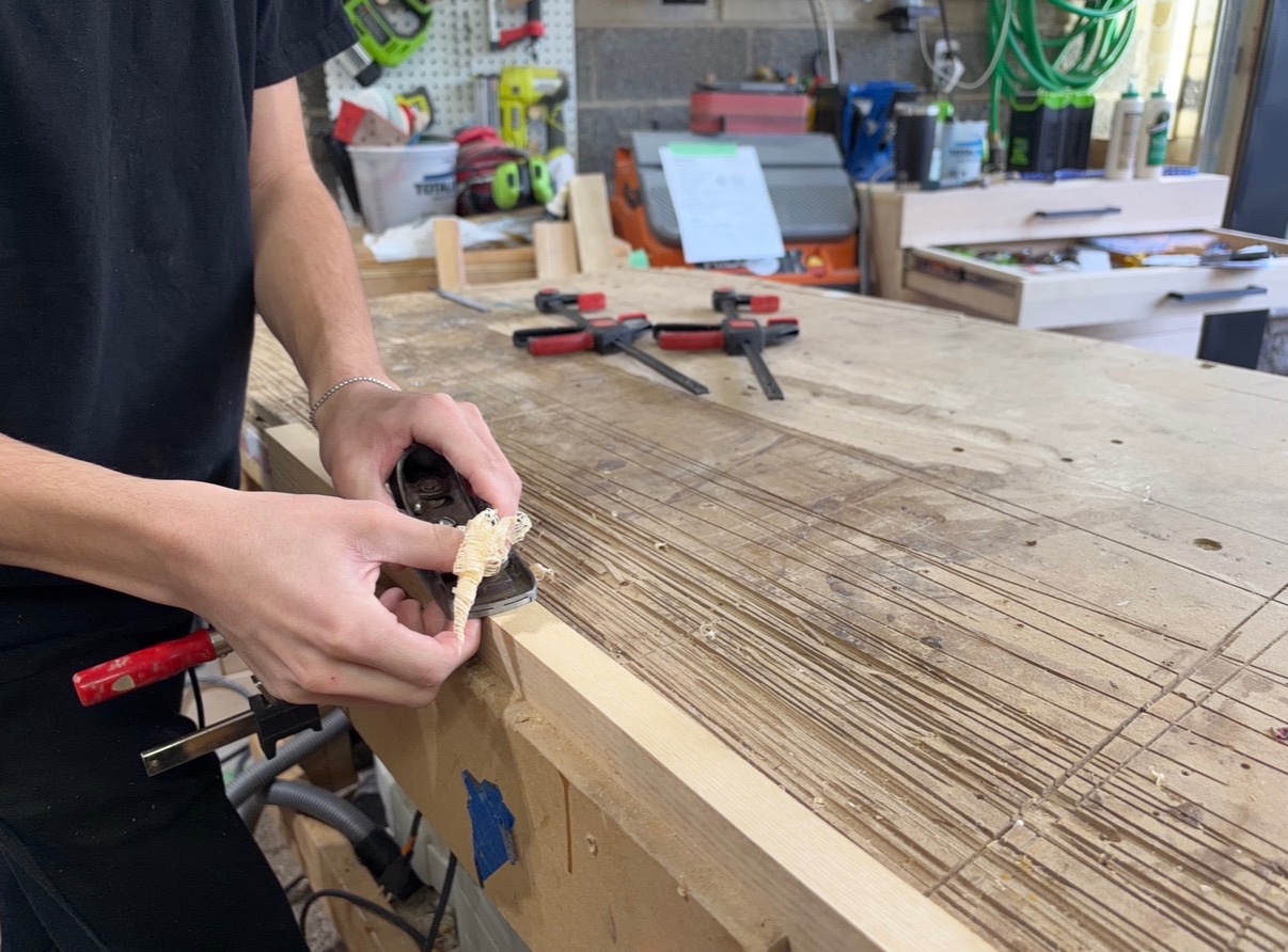Woodworker using a block plane to produce thin shavings from the edge of a secured board.
