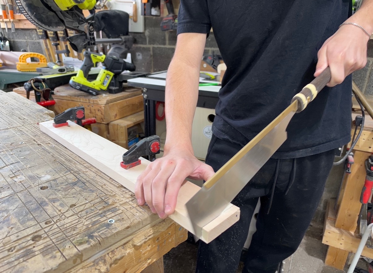 Woodworker using a Japanese pull saw to cut a clamped wooden board by hand.