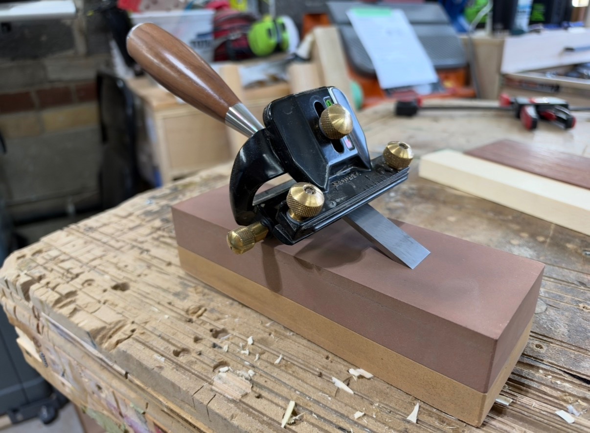Block-plane blade held in a honing guide on sharpening stones at a workshop bench.